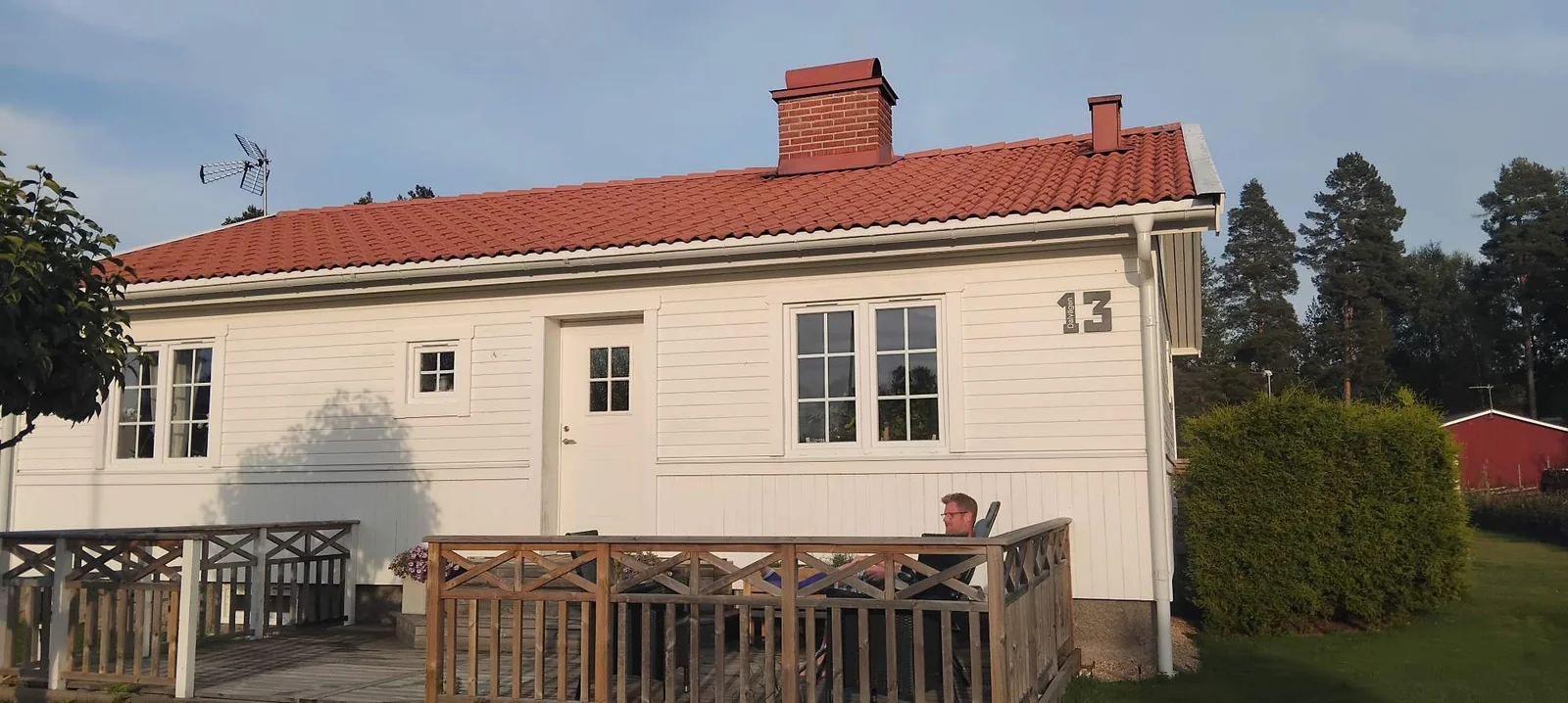 Man sitter på ett trädäck framför ett vitt trähus med rent rött tegeltak